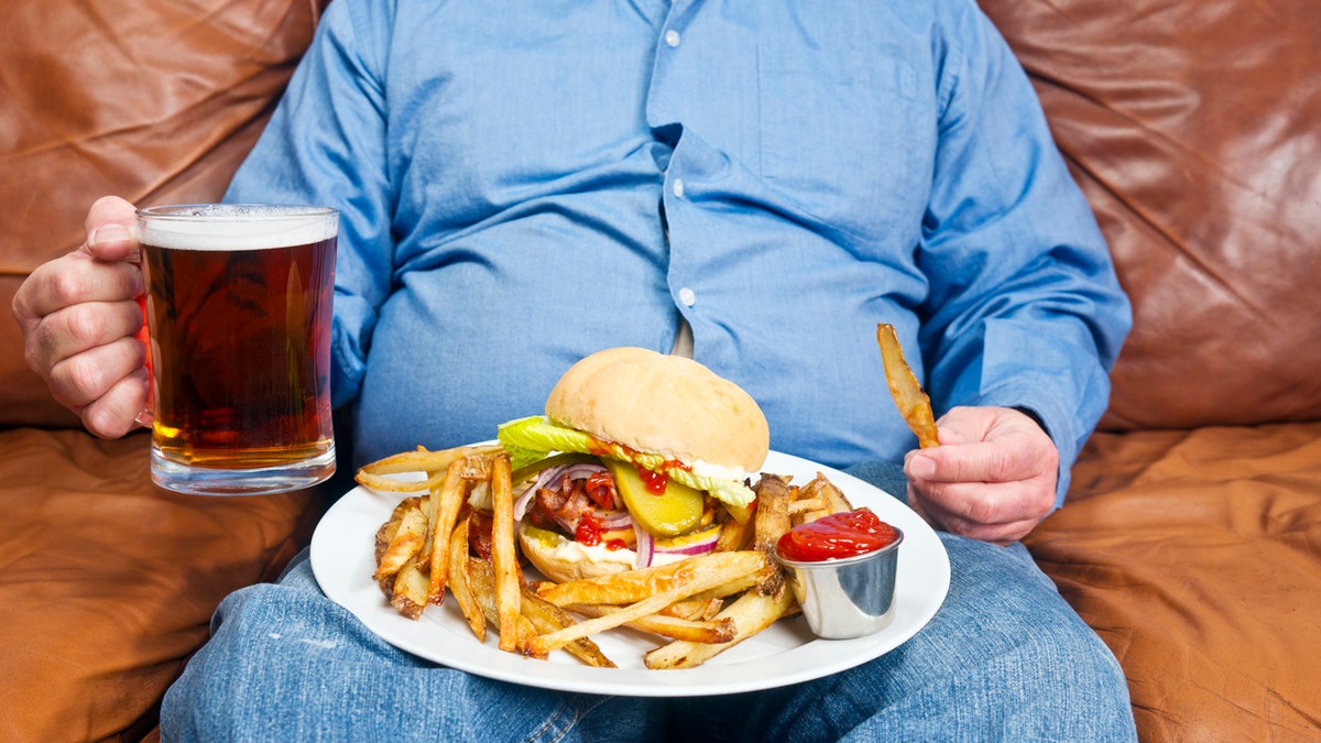 A photo of an overweight man sitting on an old sofa with a very unhealthy meal on his lap and a look of beer in his hand. Obesity is the main cause of diabetes.